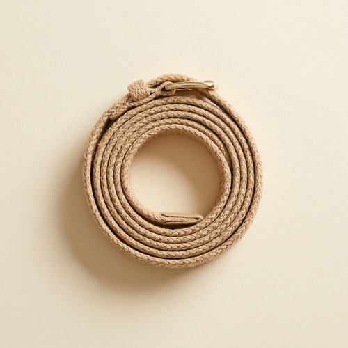 Anais sand woven belt with a gold buckle, coiled on a cream studio backdrop